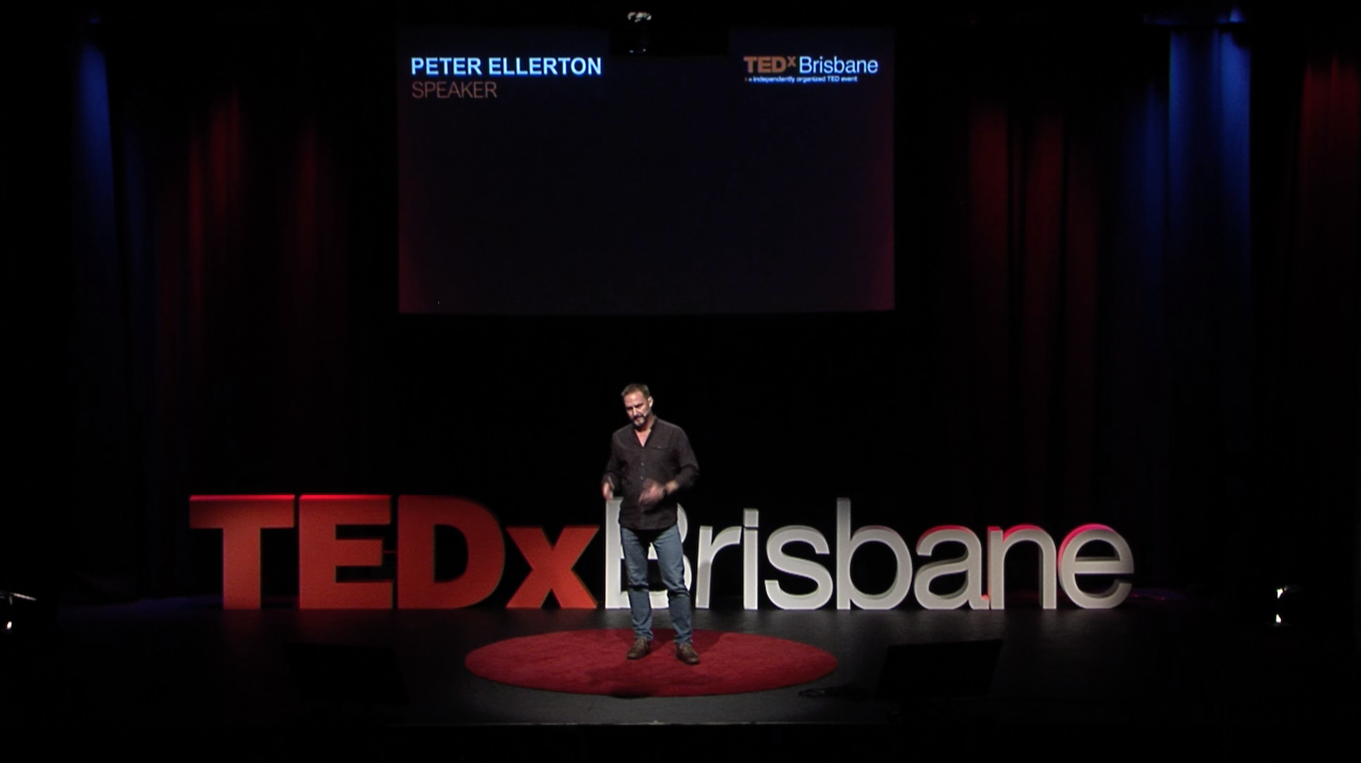 A tall man stand on a dark stage at the Tivoli Theatre, Brisbane. He stands on a small red circular carpet in front of a large TEDx Brisbane sign on the floor behind him and a projected presentation above him. His name is Peter Ellerton.