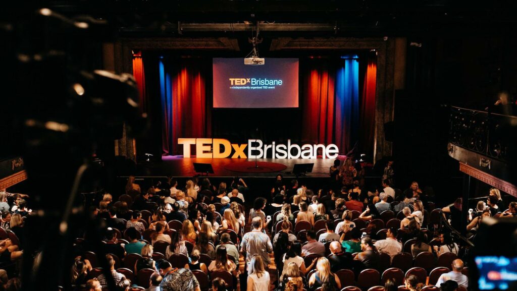 A wide shot of darkened Tivoli Theatre in Brisbane with a full audience looking at the stage that has a projector screen showing 'TEDx Brisbane.' A large sign sits on the floor of the stage, also saying 'TEDx Brisbane.' This event was live-streamed by Queensland Live Streaming