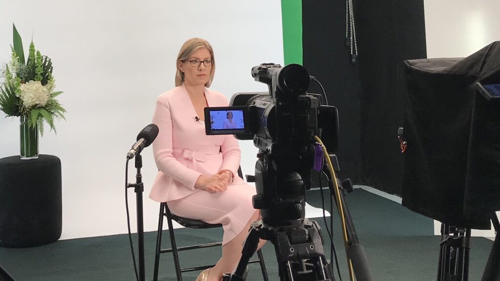 A woman sits on a chair in a video recoding studio. She is wering a pink corporate skirt and jacket suit. She has short blonde hair. There is a video camera, shotgun microphone and teleprompter in the foreground. The background is a white wall with a green screen poking out one edge. There is a short black block with a vase of tall flowers against the white wall. The floor is black in the video studio. From a production recorded by Queensland Live Streaming.